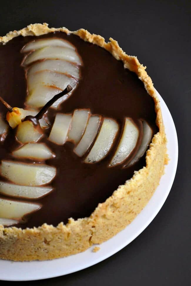 A dark chocolate pear tart with a crumbly crust, topped with sliced poached pears in a fan shape and covered in glossy ganache, is displayed on a white plate against a dark background.