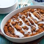 A white dish of apple cinnamon bread pudding drizzled with white icing sits on a wooden board. Nearby are stacked plates, forks, and a bowl of fresh blueberries on a turquoise table.