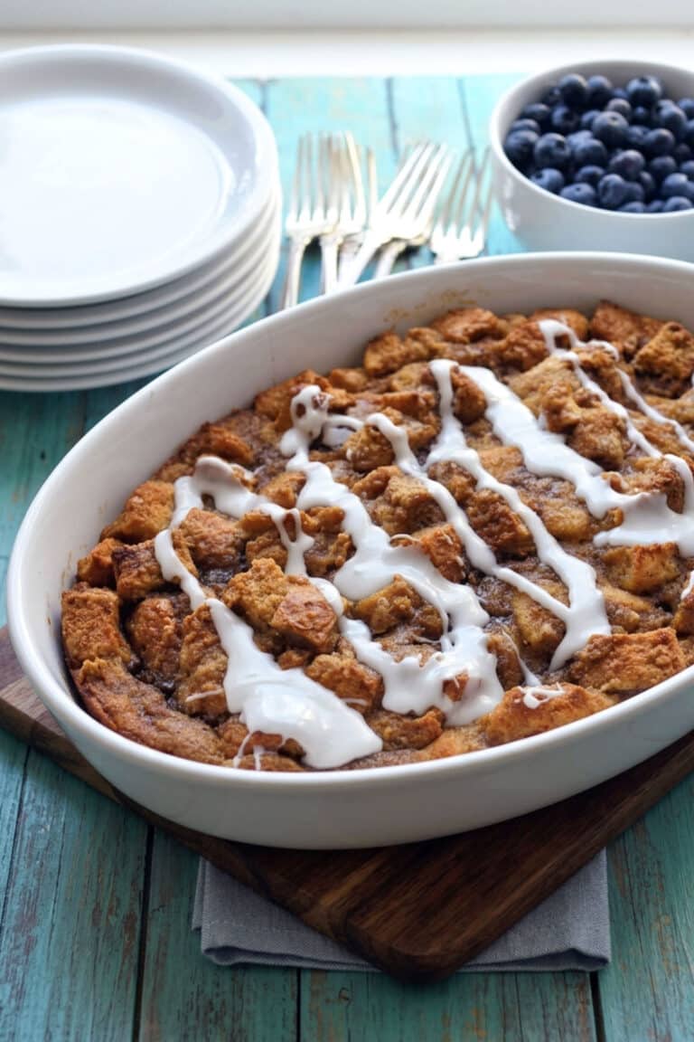 A white dish of apple cinnamon bread pudding drizzled with white icing sits on a wooden board. Nearby are stacked plates, forks, and a bowl of fresh blueberries on a turquoise table.