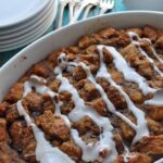 A baked apple cinnamon bread pudding drizzled with white icing sits in a white dish, with a stack of plates, metal forks, and a bowl of fresh blueberries in the background on a turquoise wooden table.