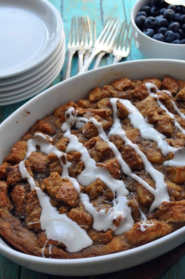 A baked apple cinnamon bread pudding drizzled with white icing sits in a white dish, with a stack of plates, metal forks, and a bowl of fresh blueberries in the background on a turquoise wooden table.