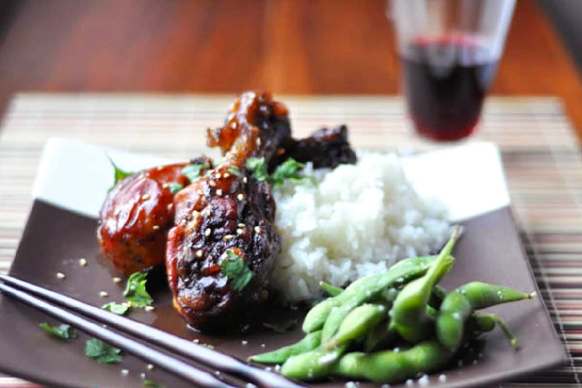 A plate with tender glazed chicken drumsticks, white rice, and steamed edamame, garnished with herbs and sesame seeds. Chopsticks rest on the side, and a glass of red beverage is blurred in the background.