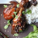 Two glazed chicken drumsticks garnished with sesame seeds and cilantro are served with a mound of white rice, a side of edamame, and chopsticks resting beside the chicken drumsticks for an authentic touch.
