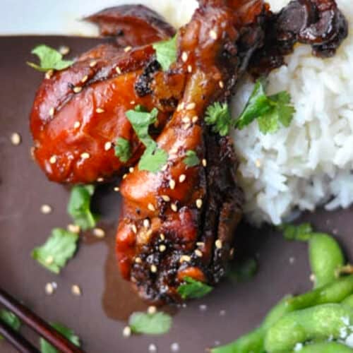 Two glazed chicken drumsticks garnished with sesame seeds and cilantro are served with a mound of white rice, a side of edamame, and chopsticks resting beside the chicken drumsticks for an authentic touch.