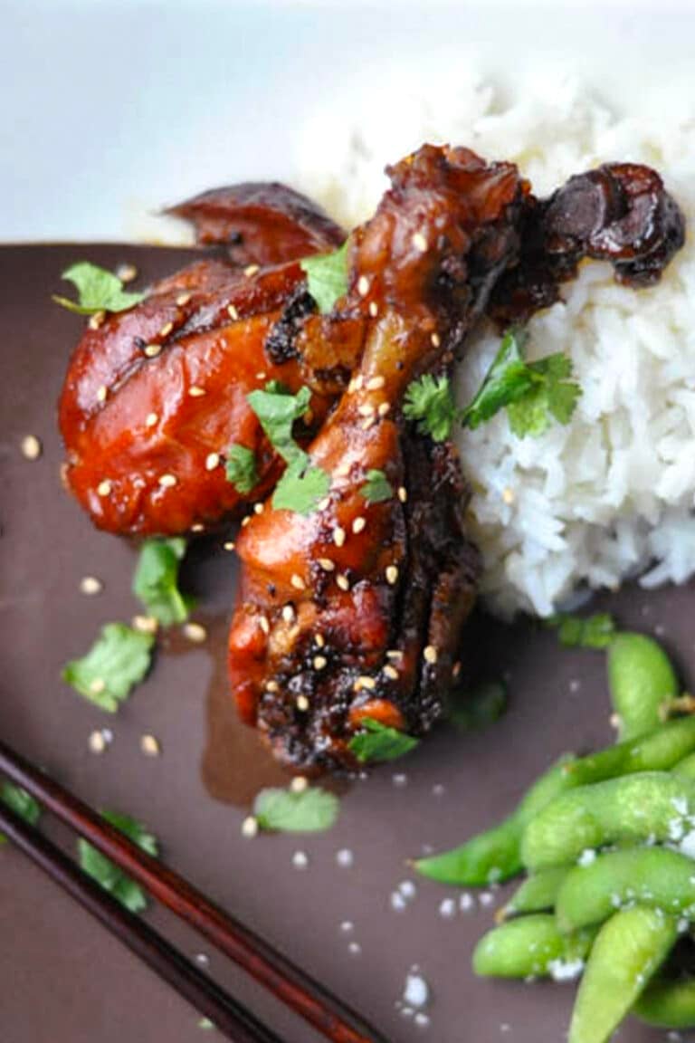 Two glazed chicken drumsticks garnished with sesame seeds and cilantro are served with a mound of white rice, a side of edamame, and chopsticks resting beside the chicken drumsticks for an authentic touch.