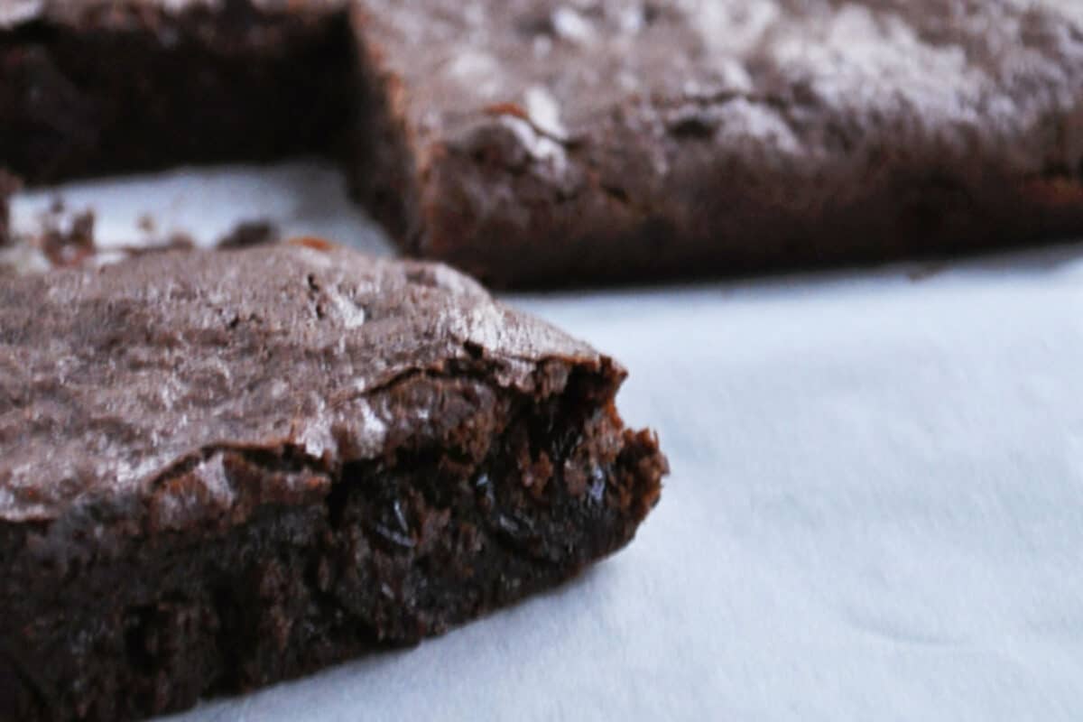A close-up of a dutch processed cocoa brownie with a bite taken out of the corner, resting on a white surface. Another uncut brownie from this chewy brownie recipe is blurred in the background.