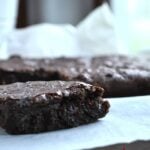 Close-up of a fudgy chocolate cherry brownie with a bite taken out, resting on white parchment paper, with more brownies in the background. Text overlay reads chocolate cherry brownies farmgirl gourmet.