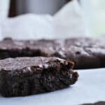 A close-up of a fudgy dark chocolate brownie with a bite taken out, resting on white parchment paper with blurred brownies in the background.