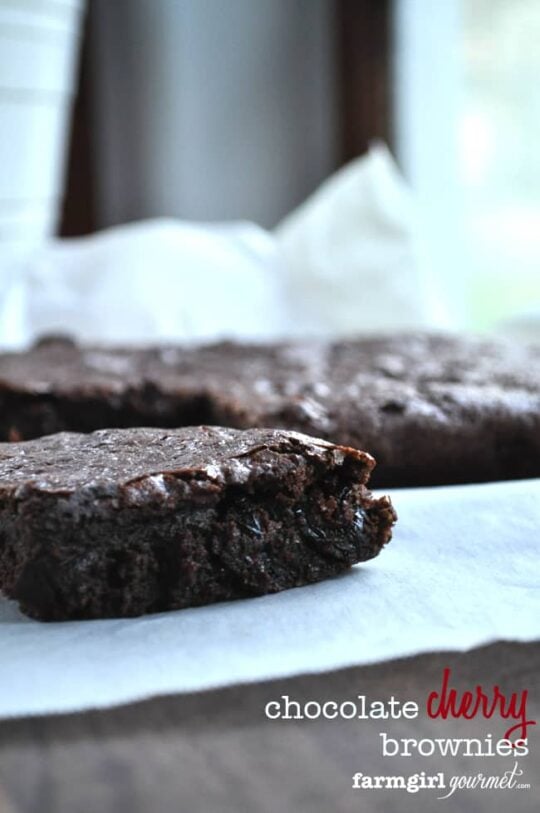 Close-up of a fudgy chocolate cherry brownie with a bite taken out, resting on white parchment paper, with more brownies in the background. Text overlay reads chocolate cherry brownies farmgirl gourmet.