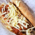 A classic meatball sub filled with juicy meatballs in tomato sauce, topped with shredded cheese and nestled in a crusty roll, served on white paper with pink polka dots. Perfect for true meatball subs fans!.