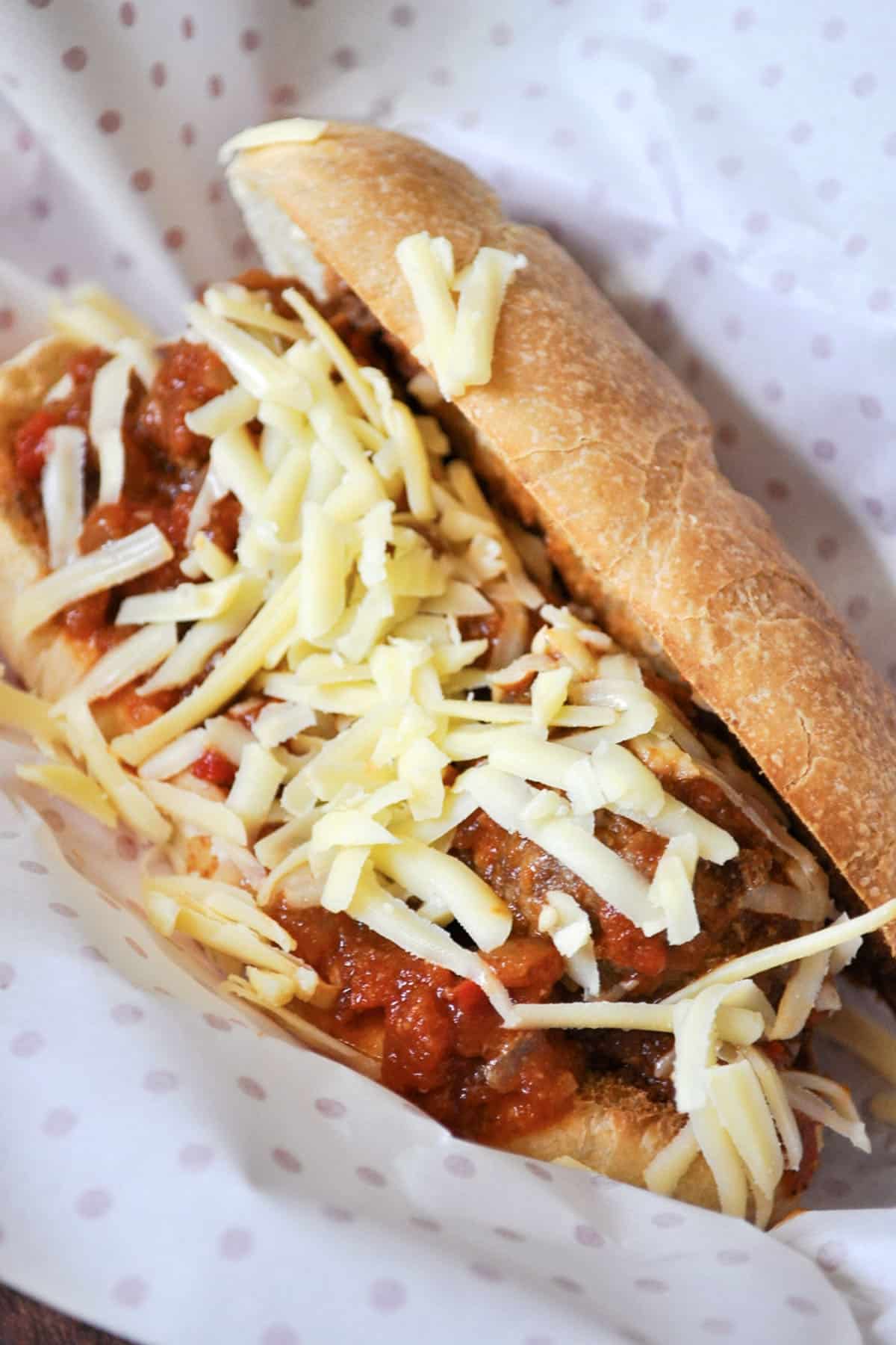 A classic meatball sub filled with juicy meatballs in tomato sauce, topped with shredded cheese and nestled in a crusty roll, served on white paper with pink polka dots. Perfect for true meatball subs fans!.