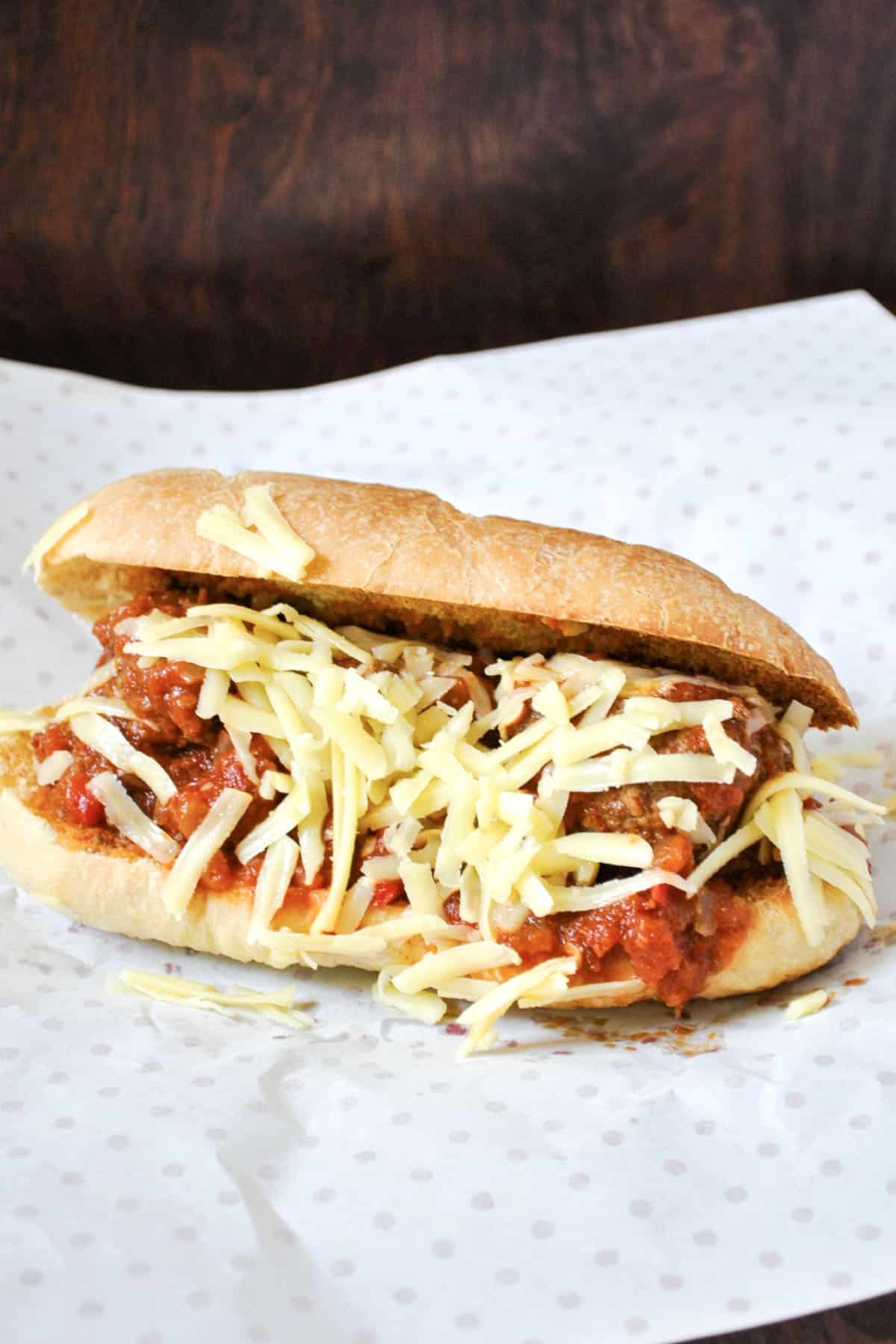 A tasty meatball sub filled with meatballs in tomato sauce and topped with shredded cheese sits on a sheet of white parchment paper with small gray dots.