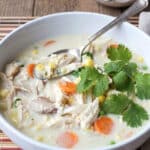 A bowl of creamy Thai chicken chowder with corn, carrots, and peas, topped with fresh cilantro, with a spoon resting inside and a side of lime wedges and chili peppers.