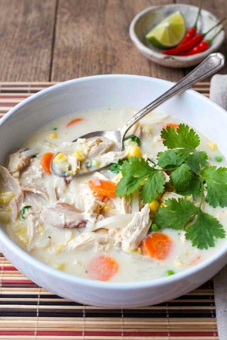 A bowl of creamy Thai chicken chowder with corn, carrots, and peas, topped with fresh cilantro, with a spoon resting inside and a side of lime wedges and chili peppers.
