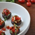 A white dish holds four mushroom bruschetta bites, stuffed and topped with chopped tomatoes, basil, and pine nuts. In the background, fresh basil leaves and grape tomatoes rest on a wooden surface.