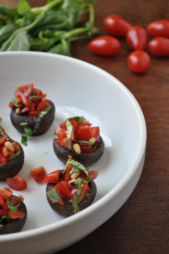 A white dish holds four mushroom bruschetta bites, stuffed and topped with chopped tomatoes, basil, and pine nuts. In the background, fresh basil leaves and grape tomatoes rest on a wooden surface.