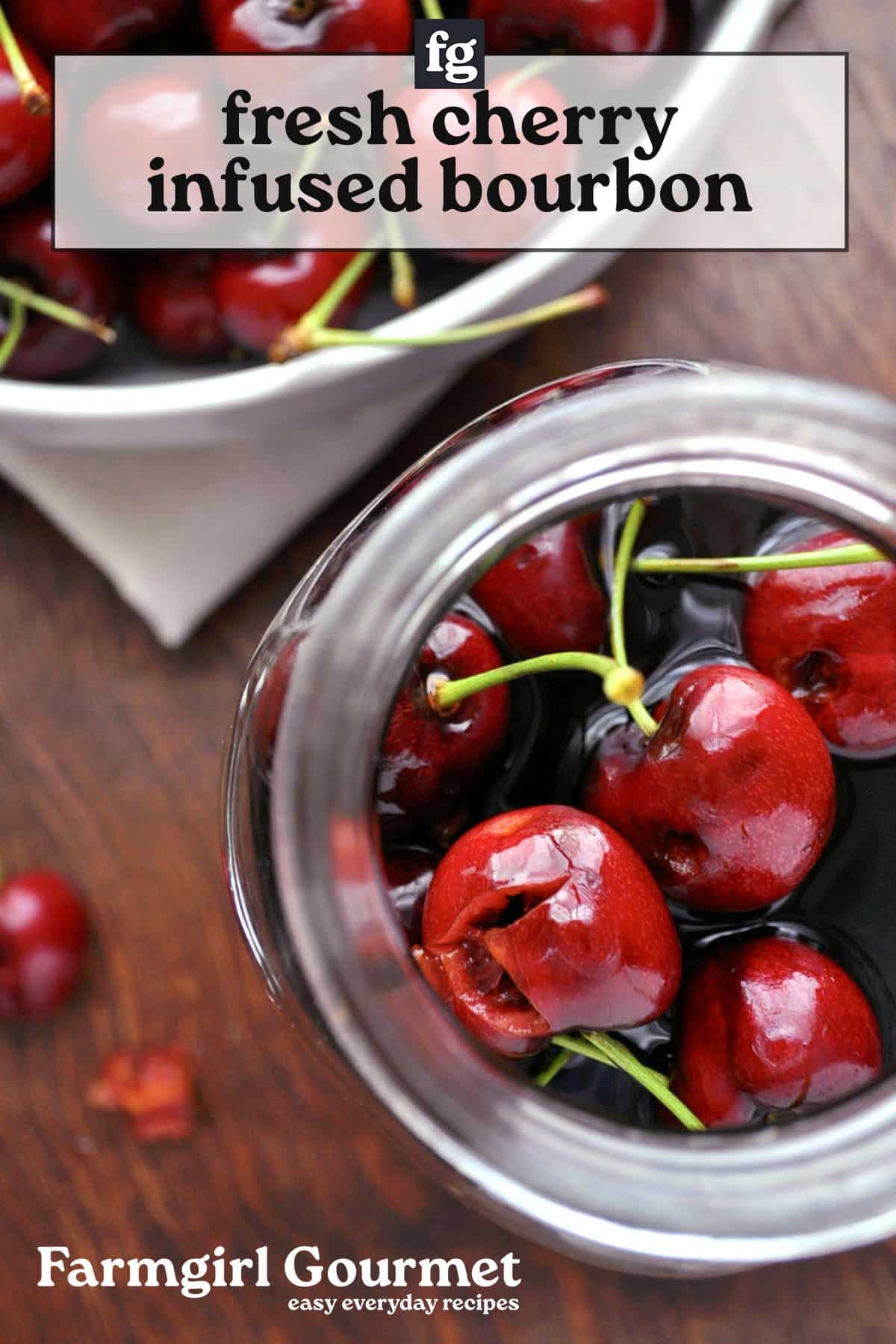 A jar filled with fresh cherries and cherry bourbon sits on a wooden surface, next to a white bowl of cherries. Text reads fresh cherry infused bourbon and Farmgirl Gourmet easy everyday recipes.