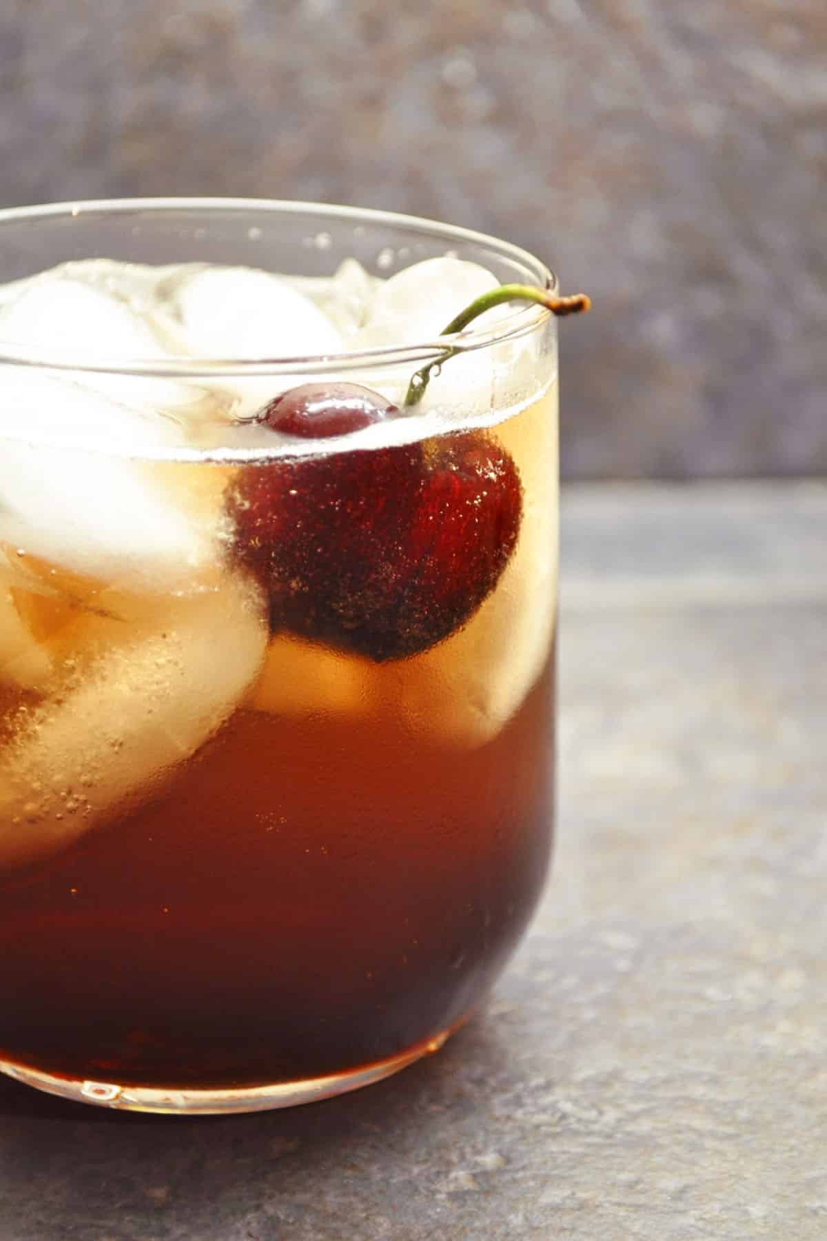 A glass of dark soda filled with ice cubes and a fresh cherry bourbon garnish on top, set against a neutral, textured background.