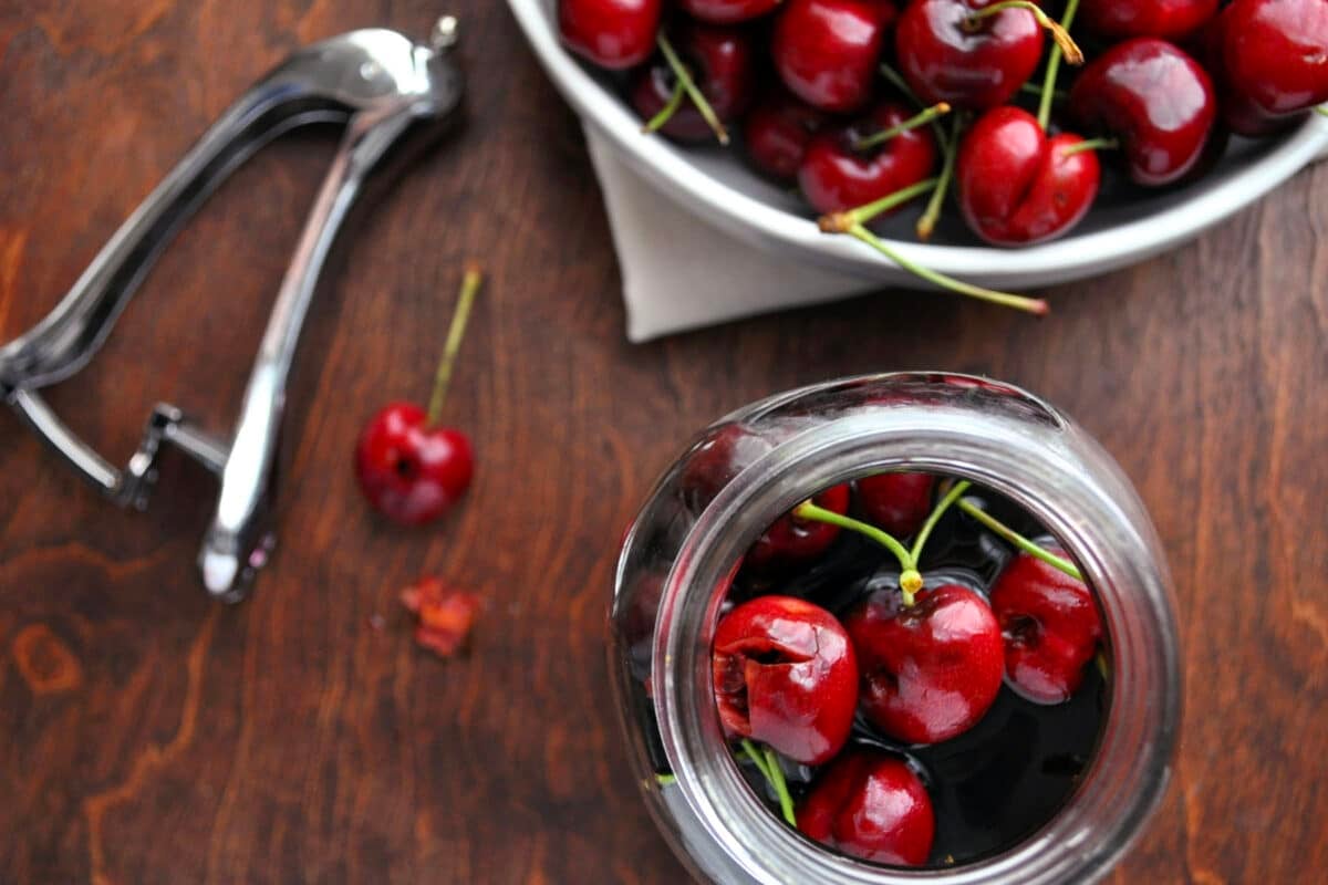 A jar filled with fresh cherries sits on a wooden surface, next to a bowl of cherries, a metal cherry pitter, and a splash of cherry bourbon. Two cherries and a cherry stem rest on the table.