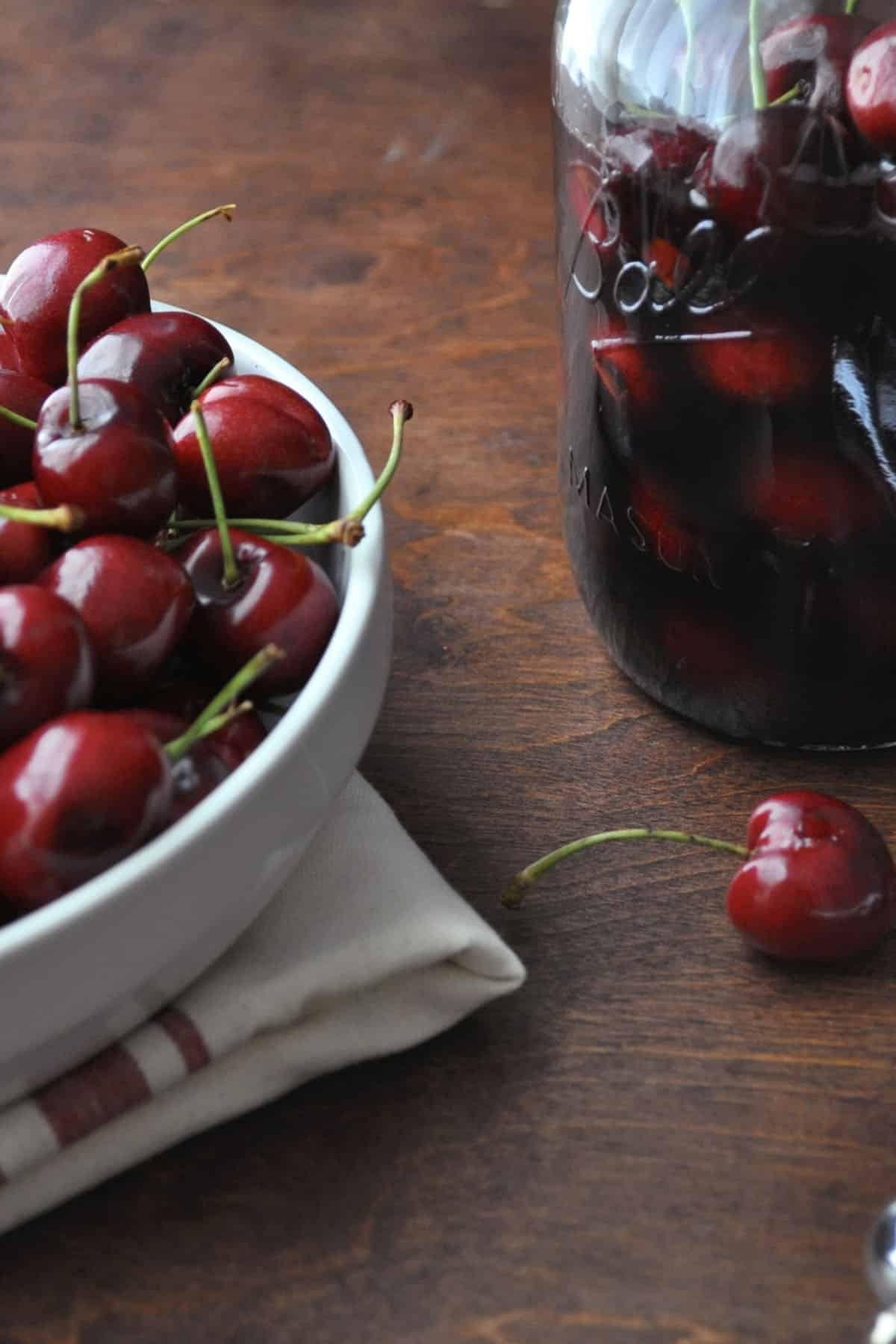 A white bowl filled with fresh cherries sits on a folded cloth next to a glass jar of cherry bourbon preserves on a wooden surface; a few loose cherries are scattered nearby.