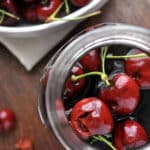 A close-up top view of bright red cherries with stems inside a glass jar of cherry bourbon, with more cherries in a bowl in the background on a wooden surface.