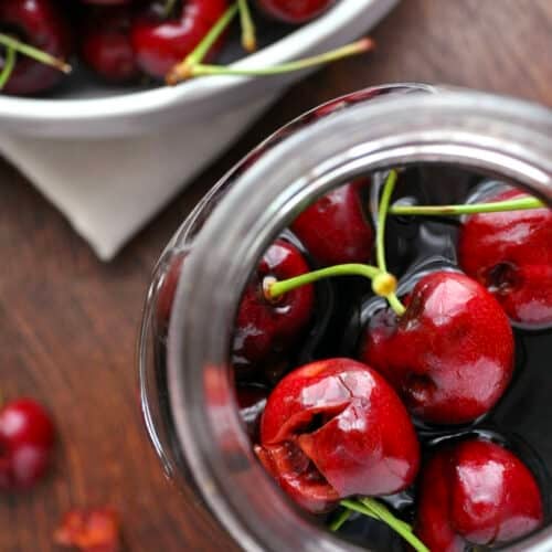 A close-up top view of bright red cherries with stems inside a glass jar of cherry bourbon, with more cherries in a bowl in the background on a wooden surface.