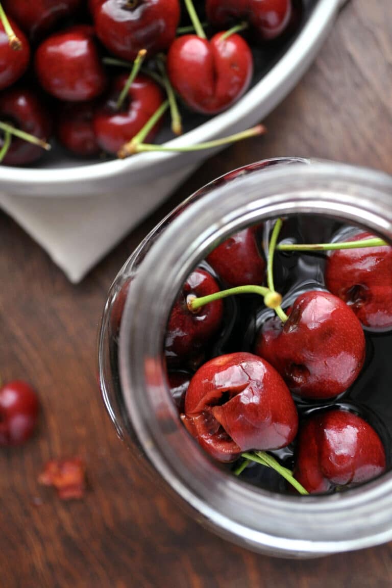 A close-up top view of bright red cherries with stems inside a glass jar of cherry bourbon, with more cherries in a bowl in the background on a wooden surface.