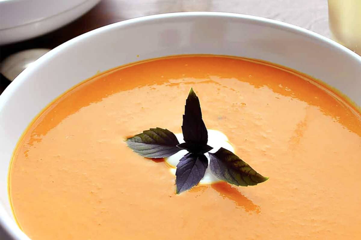 A bowl of creamy chipotle tomato soup garnished with a dollop of cream and three basil leaves arranged in a star shape at the center.