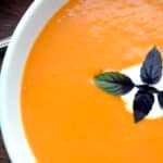 A close-up of creamy chipotle tomato soup in a white bowl, topped with a swirl of cream and a sprig of fresh basil.