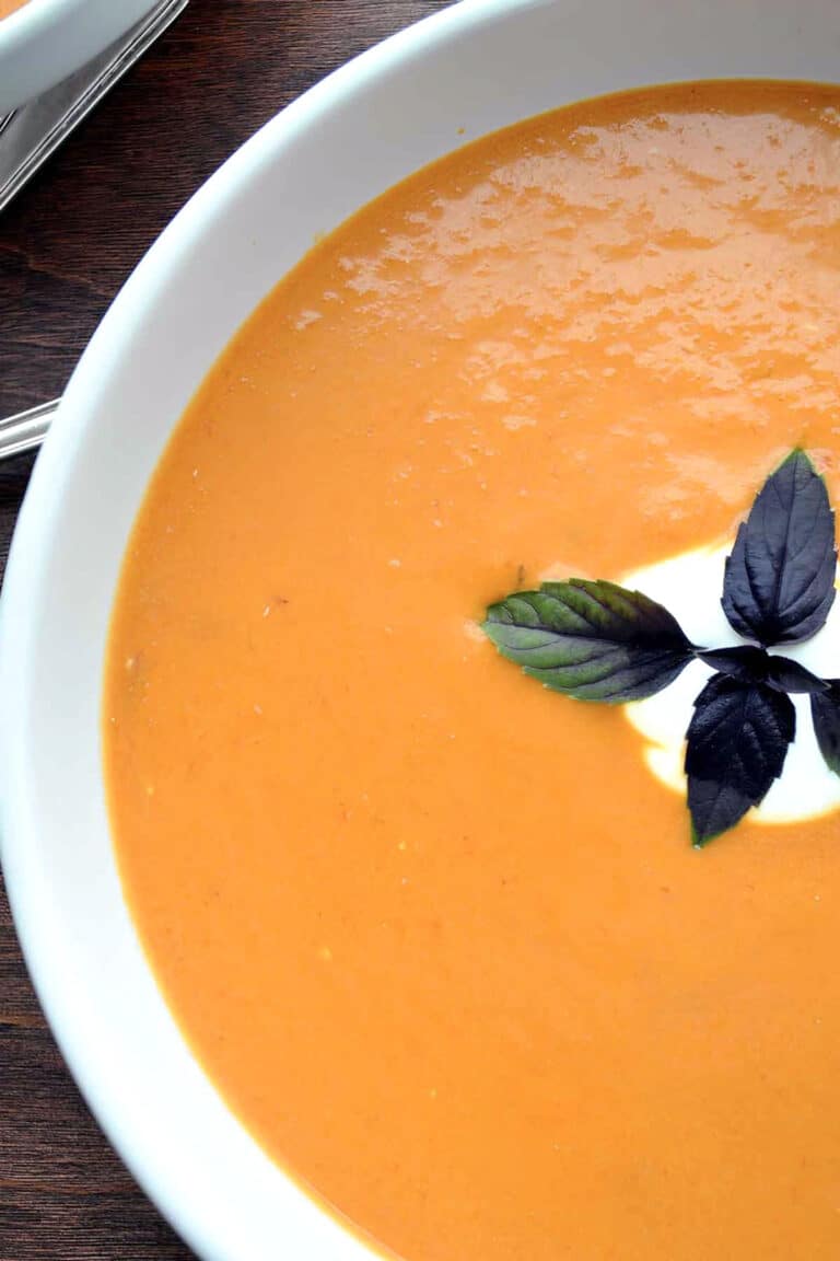 A close-up of creamy chipotle tomato soup in a white bowl, topped with a swirl of cream and a sprig of fresh basil.