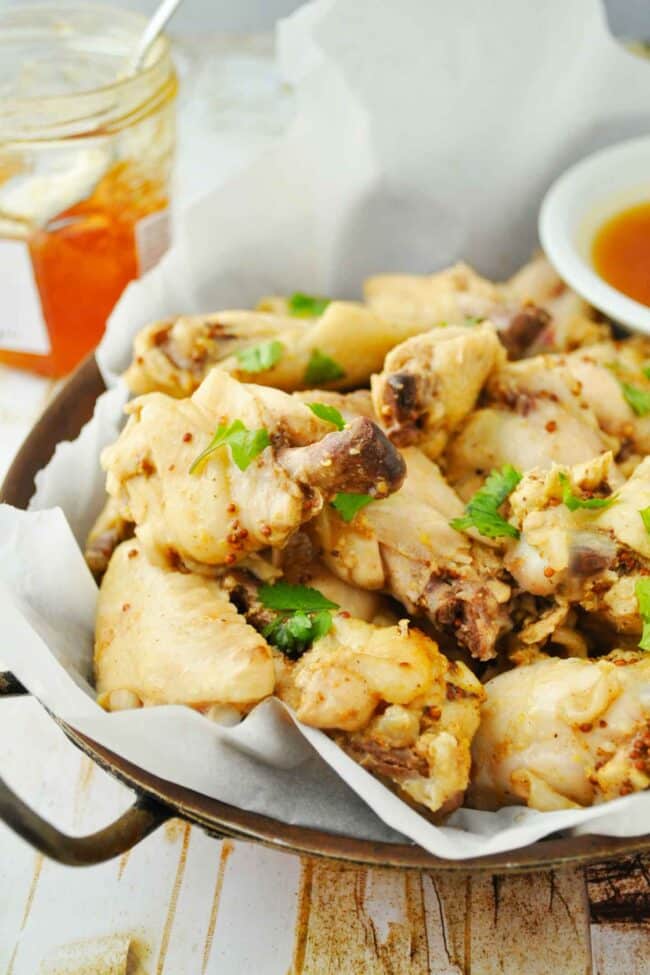 A tray of mustard glazed chicken wings garnished with fresh herbs sits on parchment paper, with a small bowl of dipping sauce and a jar of honey in the background.
