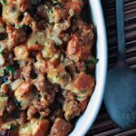 Casserole dish filled with sweet bread stuffing made of sausage, bread cubes, onions, and herbs, resting on a striped placemat with a metal serving spoon beside it.