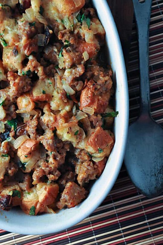 Casserole dish filled with sweet bread stuffing made of sausage, bread cubes, onions, and herbs, resting on a striped placemat with a metal serving spoon beside it.
