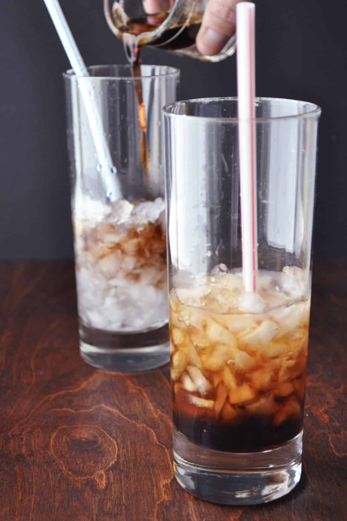 Two tall glasses filled with ice and a straw each, while a hand pours a dark liquid—perhaps a classic Kahlúa and Coke—into the glass in the foreground. The scene is set on a dark wooden surface with a black background.