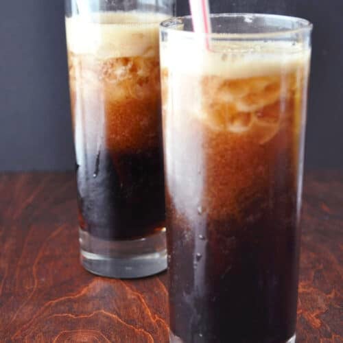 Two tall glasses of Kahlúa and coke with ice sit on a wooden surface. Each glass has a straw—one blue, one pink. The drinks are dark and fizzy, with a layer of foam on top and condensation clinging to the sides.