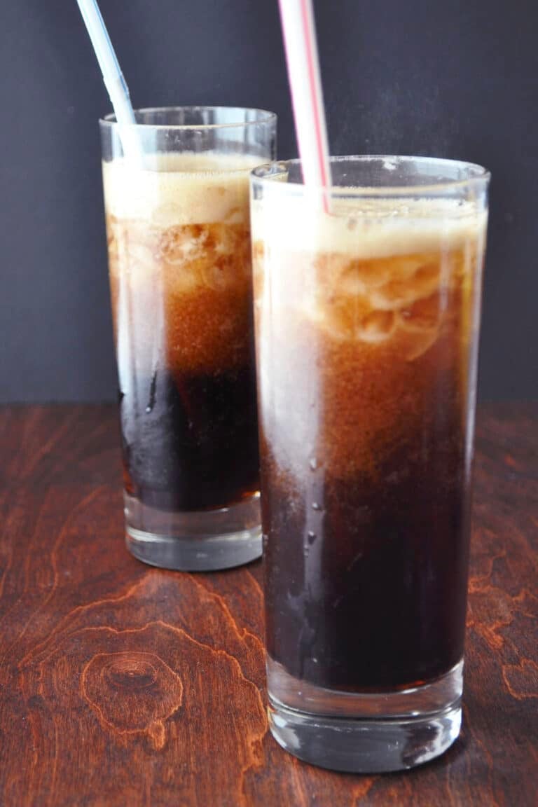 Two tall glasses of Kahlúa and coke with ice sit on a wooden surface. Each glass has a straw—one blue, one pink. The drinks are dark and fizzy, with a layer of foam on top and condensation clinging to the sides.