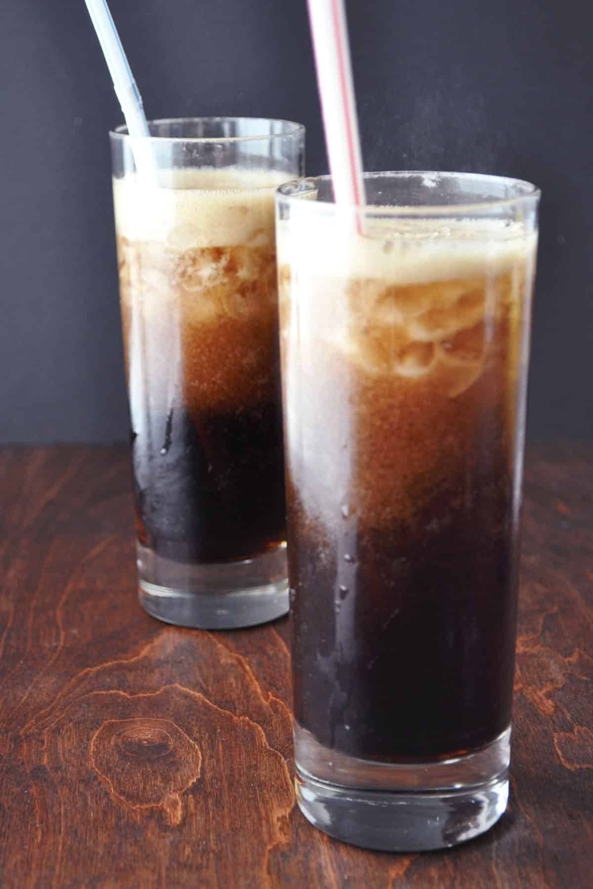 Two tall glasses of Kahlúa and coke with ice sit on a wooden surface. Each glass has a straw—one blue, one pink. The drinks are dark and fizzy, with a layer of foam on top and condensation clinging to the sides.
