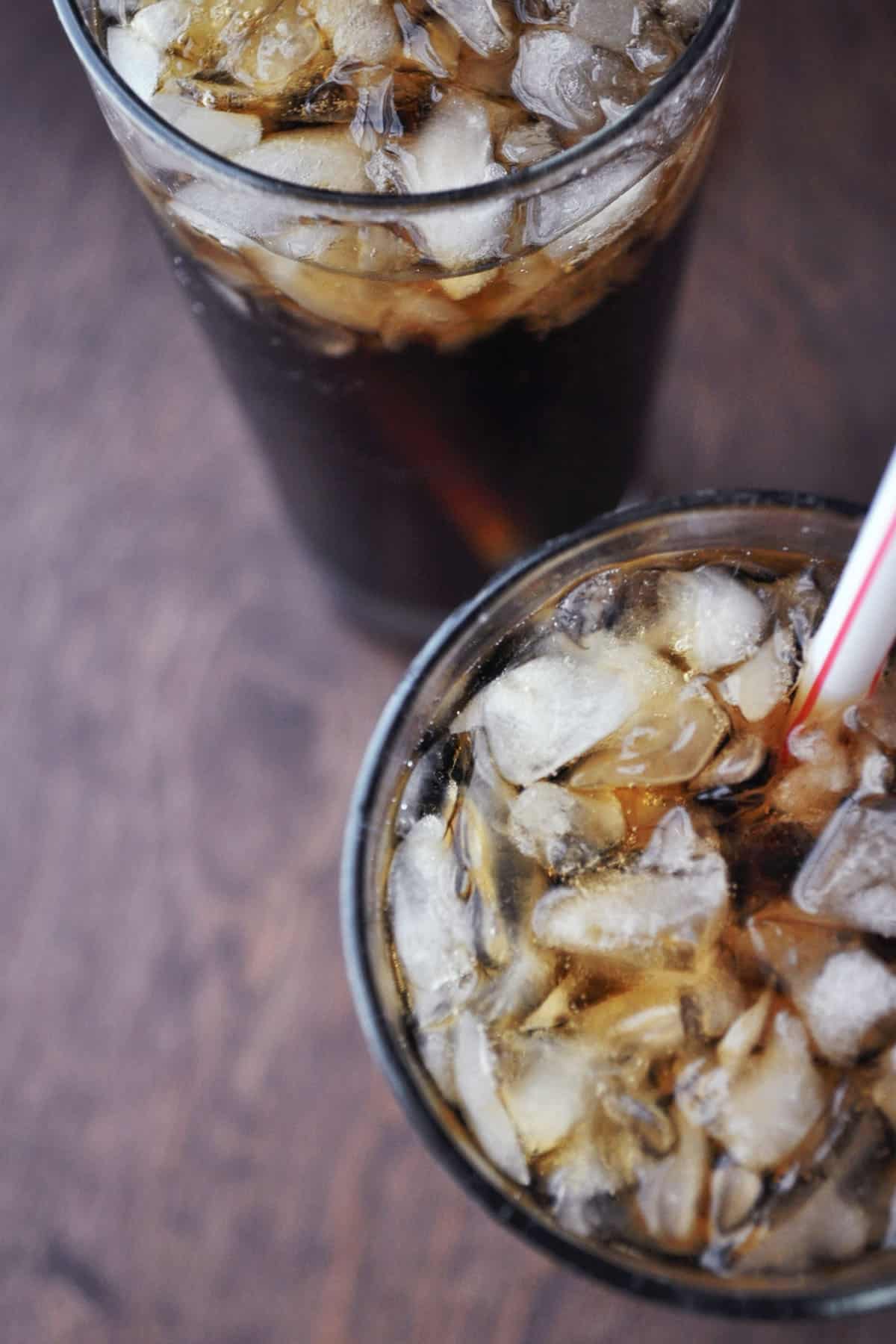 Two glasses of iced Kahlúa and Coke with red and white striped straws sit on a wooden surface. The glasses are filled with ice, and the drinks have a dark, fizzy appearance.