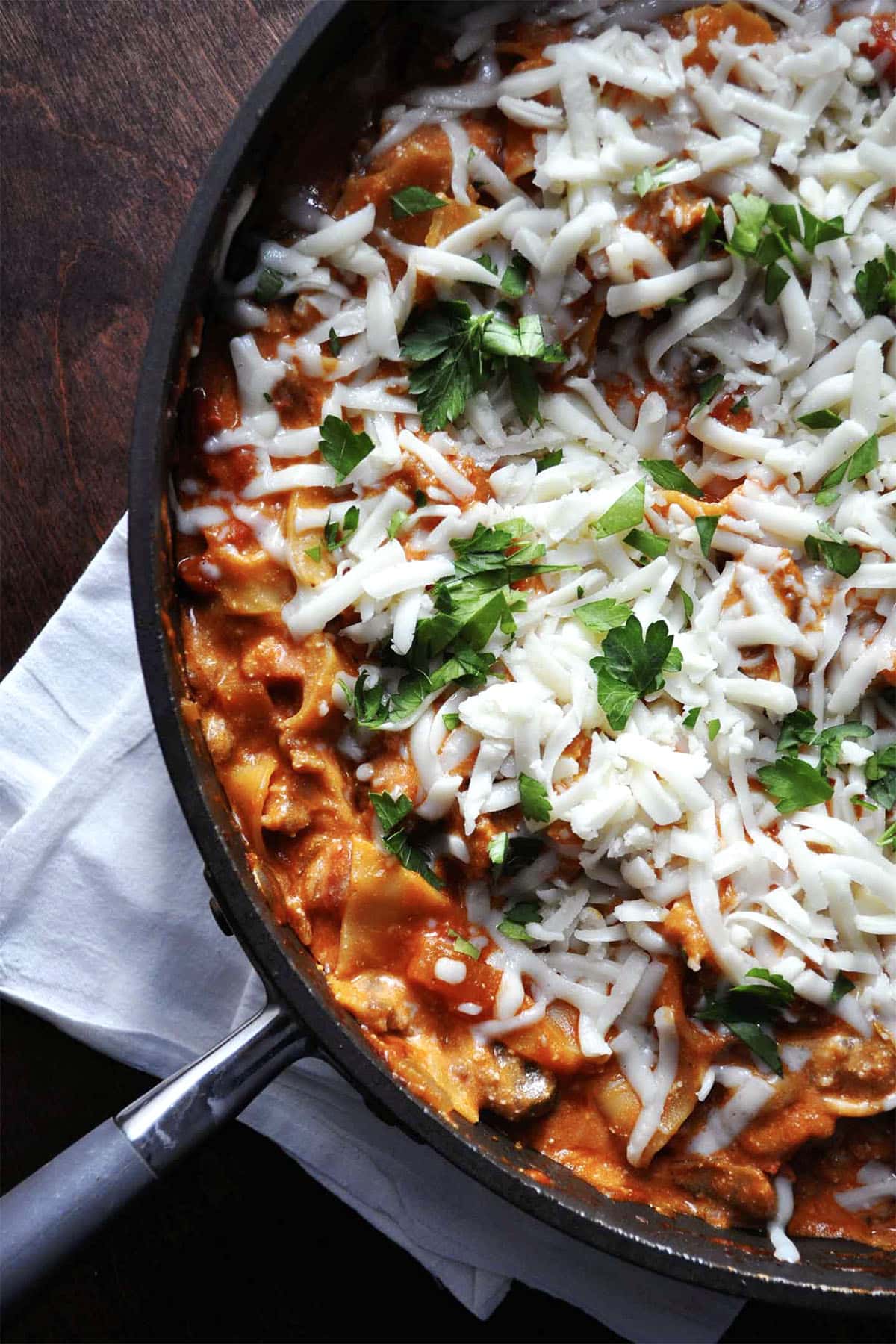 A skillet filled with cheesy skillet lasagna, topped with shredded mozzarella and fresh parsley, rests on a white napkin on a wooden surface.
