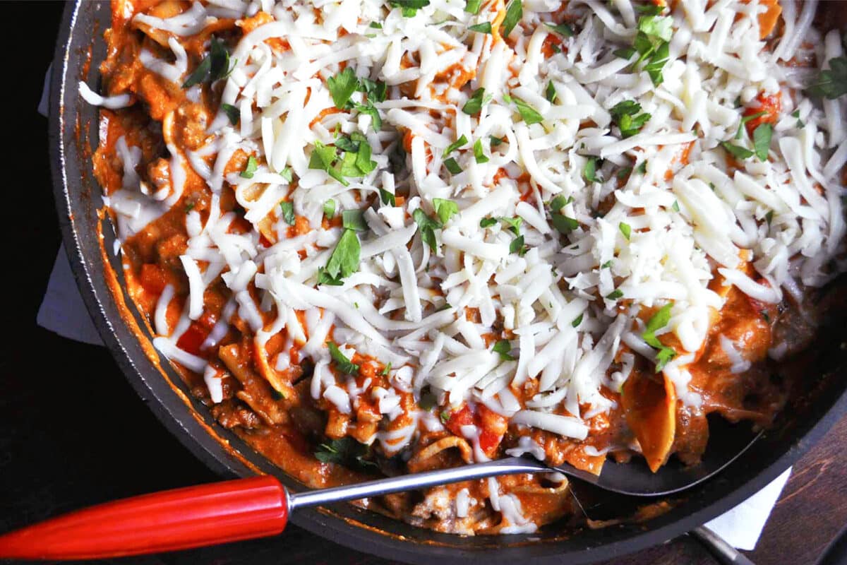 Close-up of a skillet filled with skillet lasagna in a tomato-based sauce, topped with shredded cheese and garnished with chopped fresh parsley. A serving spoon with a red handle rests in the skillet.