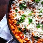A skillet of cheesy skillet lasagna topped with melted cheese and fresh parsley, resting on a white napkin on a dark wooden table.