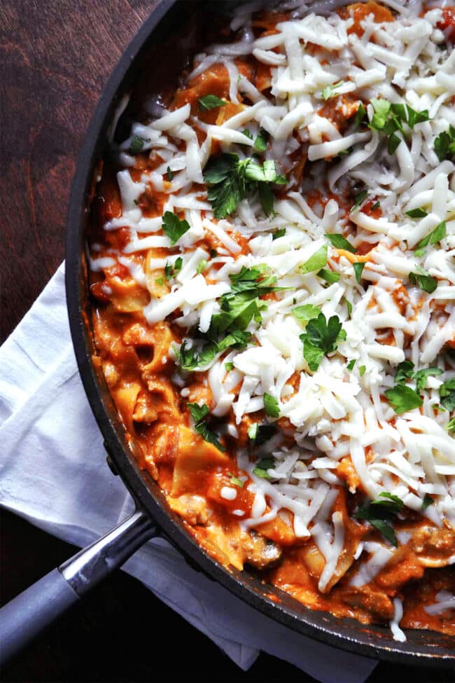 A skillet of cheesy skillet lasagna topped with melted cheese and fresh parsley, resting on a white napkin on a dark wooden table.