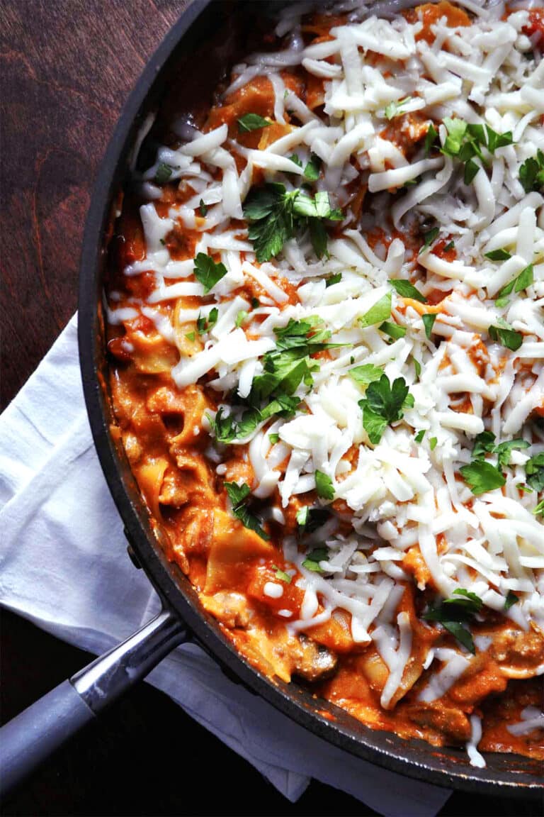A skillet of cheesy skillet lasagna topped with melted cheese and fresh parsley, resting on a white napkin on a dark wooden table.