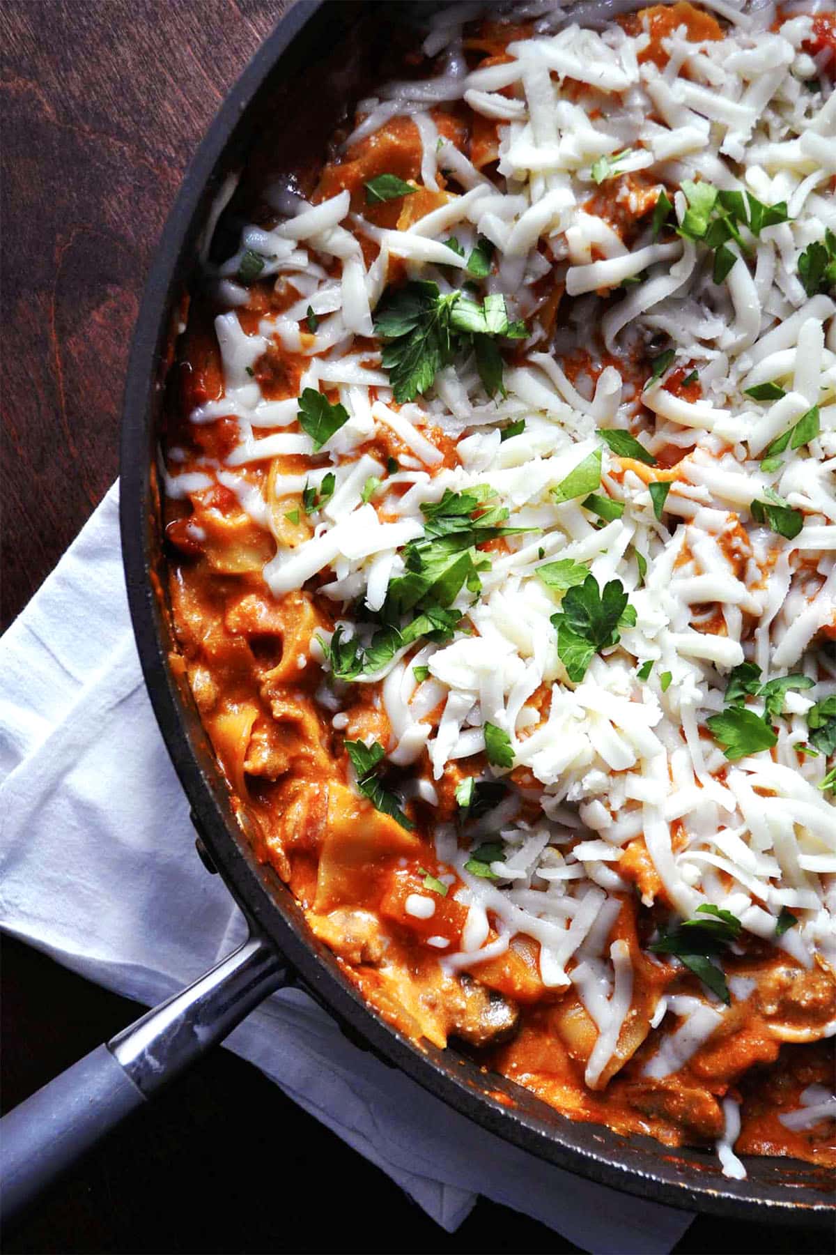 A skillet of cheesy skillet lasagna topped with melted cheese and fresh parsley, resting on a white napkin on a dark wooden table.
