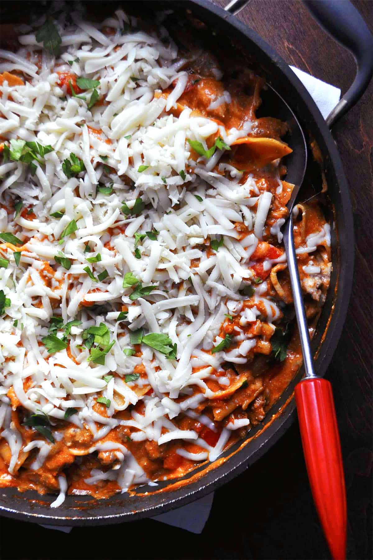 A skillet filled with skillet lasagna, topped with melted cheese and parsley, partially served with a spoon that has a red handle.