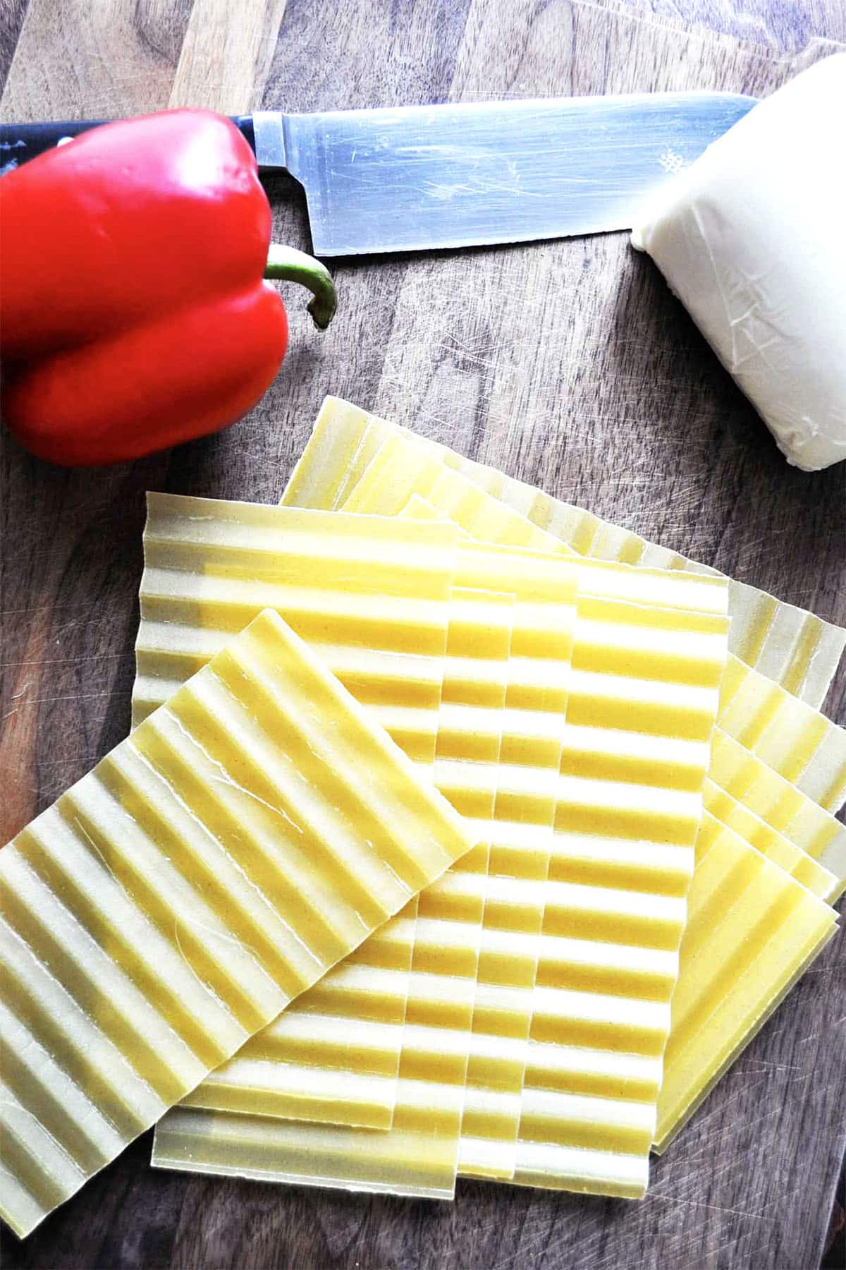 A stack of uncooked lasagna noodles on a wooden surface, next to a red bell pepper, a block of cheese, and a large kitchen knife—perfect ingredients for making skillet lasagna.
