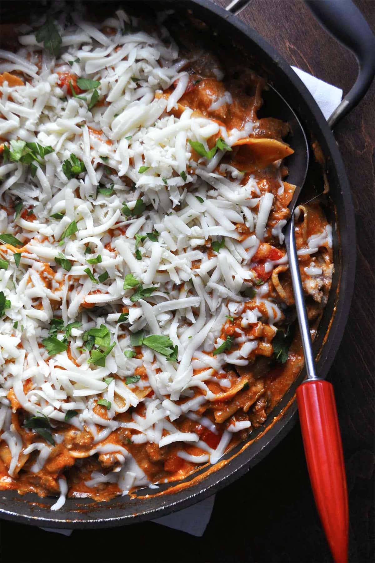 A skillet filled with skillet lasagna, topped with melted shredded cheese and chopped parsley, with a red-handled serving spoon on the side.