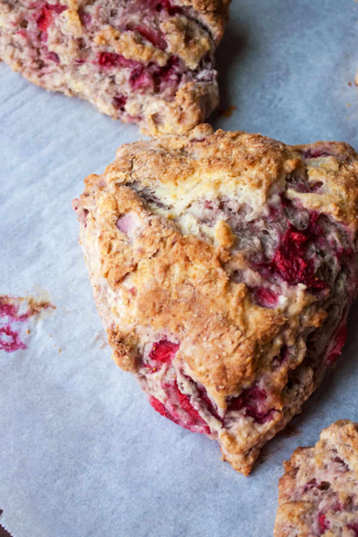 Heart-shaped scone with visible pieces of red fruit, likely fresh strawberry scones, on a piece of parchment paper.