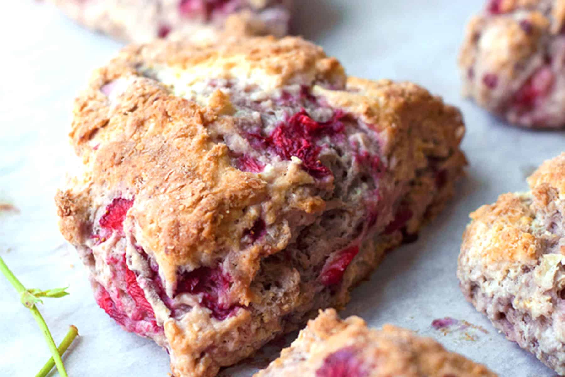 A close-up of a golden-brown fresh strawberry scone filled with visible red raspberry pieces, resting on parchment paper with other scones partially visible in the background.