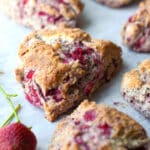 A close-up of fresh strawberry scones on parchment paper, with visible chunks of strawberries inside and a couple of whole strawberries nearby.
